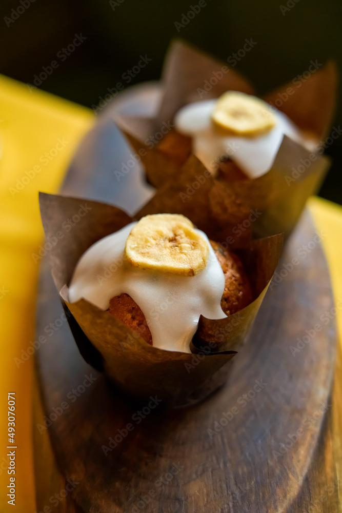 Delicious and fresh banana muffin with sugar glaze and banana chips wrapped in paper on a wooden background. Close-up. Selective focus. Top view. Blurred soft background. Delicious and fresh banana muffin with sugar glaze and banana chips wrapped in paper on a wooden background. Close-up. Selective focus. Top view. Blurred soft background.