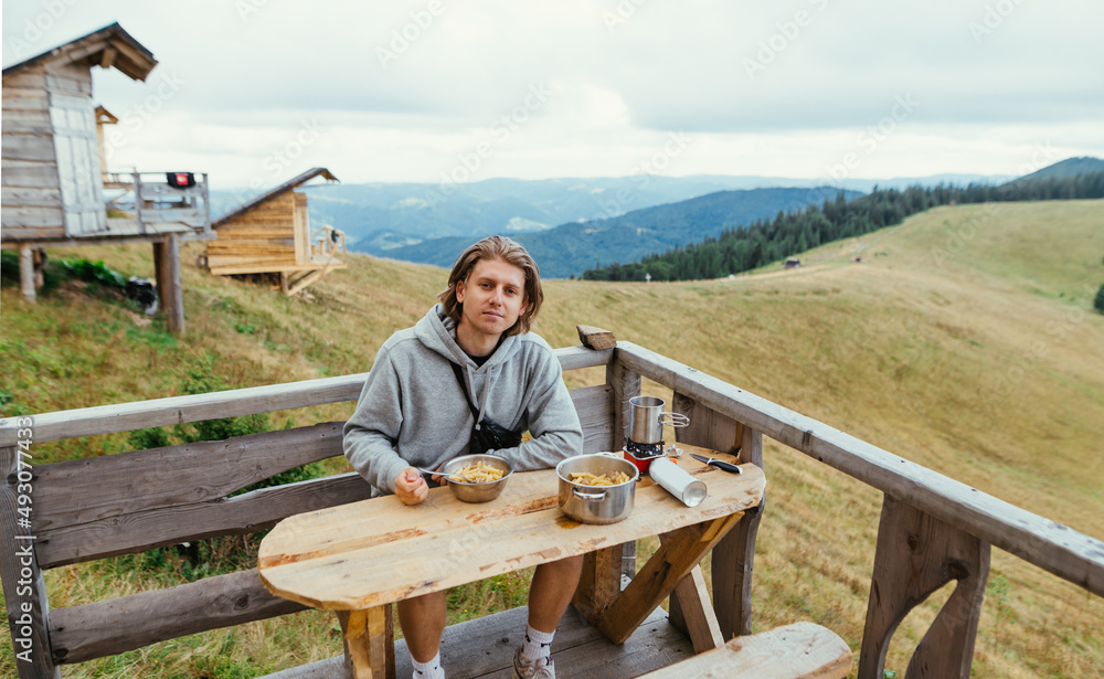 Male tourist sits at a table on the terrace of a house in the mountains and eats with a smile on his face, looking at the camera on a background of natural views and bungalows