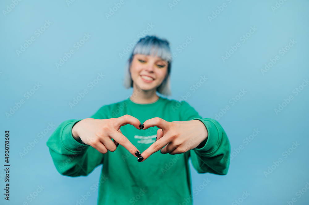 © bodnarphoto - Happy lady with colored hair with a smile on her face shows a heart gesture to the camera on a blue background. © bodnarphoto - Happy lady with colored hair with a smile on her face shows a heart gesture to the camera on a blue background.