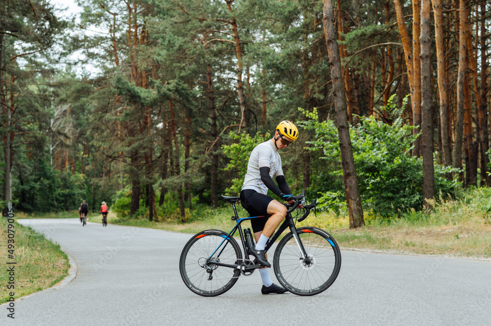 Photo of a professional cyclist in full sports gear stands on an asphalt road in the woods and poses for the camera.