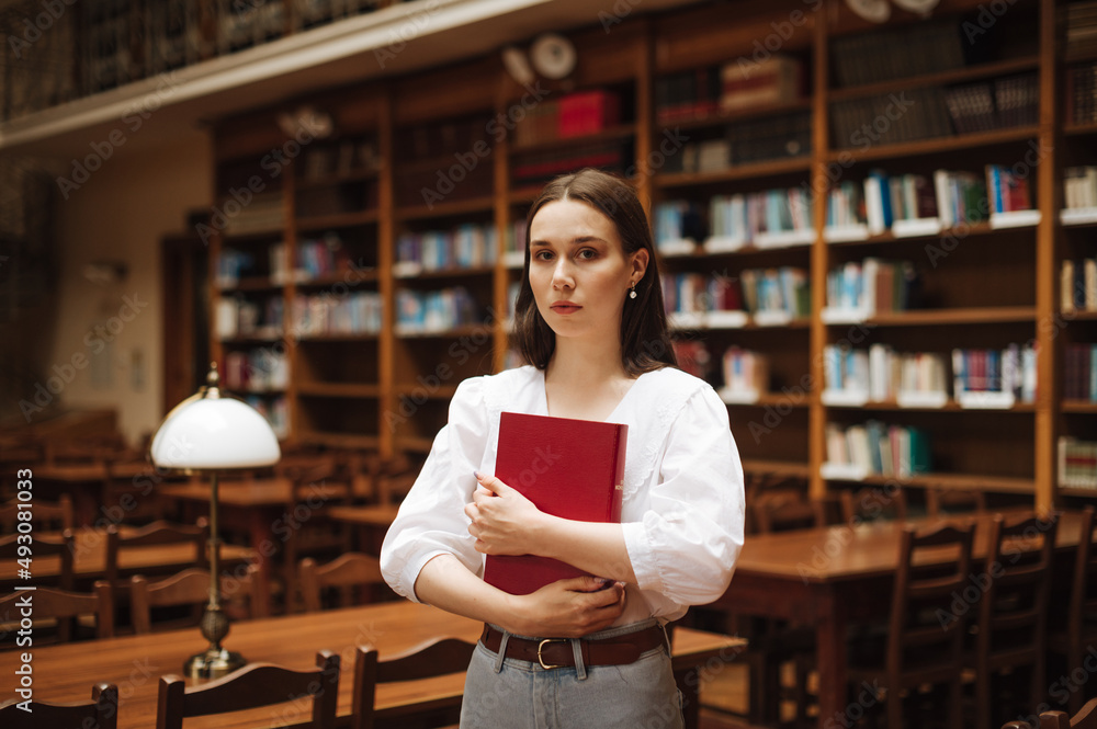 Beautiful woman in a white blouse stands with an atmospheric cozy library with a book in her hands and poses for the camera with a serious face. Beautiful woman in a white blouse stands with an atmospheric cozy library with a book in her hands and poses for the camera with a serious face.