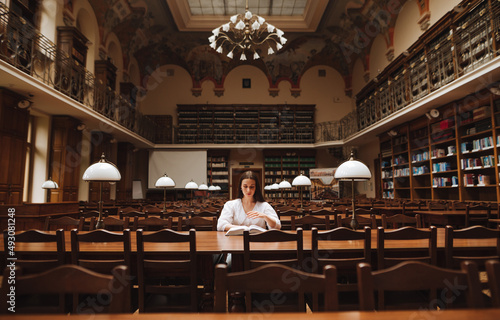 Female student sitting at a table in a beautiful old library and reading a book with a serious face