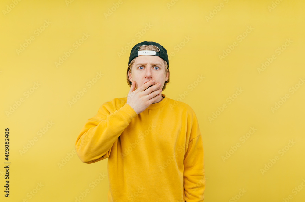 Shocked young man in a yellow sweatshirt and cap stands on a yellow background and looks at the camera with a surprised face covering his mouth with his hand. Shocked young man in a yellow sweatshirt and cap stands on a yellow background and looks at the camera with a surprised face covering his mouth with his hand.