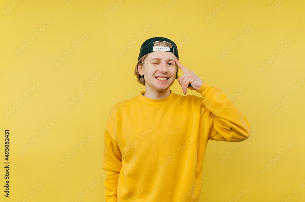 Positive young man in a cap stands on a yellow background, shows a finger on his temple showing a gesture of Positive young man in a cap stands on a yellow background, shows a finger on his temple showing a gesture of