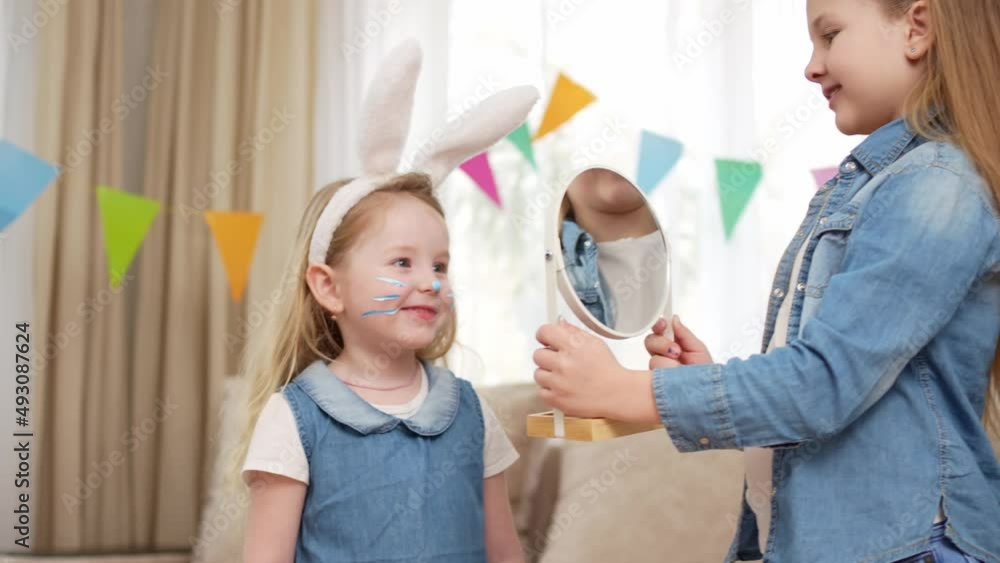 a little girl shows younger sister reflection in a mirror with a painted face. a little girl shows younger sister reflection in a mirror with a painted face.