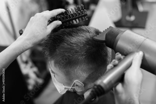 The hairdresser dries and styles the hair with the help of a hair dryer and a comb for a schoolboy boy, visiting the hairdresser during a coronavirus pandemic.
