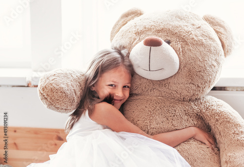 Tableau sur toile little smiling cute girl in beautiful white dress is sitting with big teddy bear