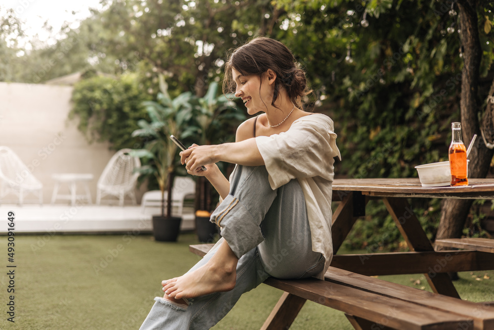 Portrait of beautiful young caucasian woman smiling while sitting on bench with mobile phone. Dark-haired model in casual light clothing. Social media addiction concept Portrait of beautiful young caucasian woman smiling while sitting on bench with mobile phone. Dark-haired model in casual light clothing. Social media addiction concept