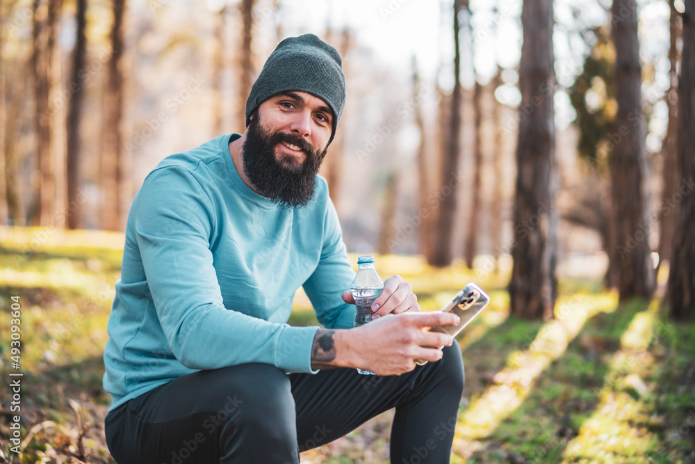 Fototapeta premium Man using phone and drinking water after exercise in the nature.