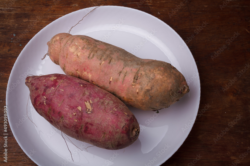 Orange, purple raw sweet potatoes, yam on white plate, wooden rustic table. Farm harvest, organic vegetables, shop local vegan food Orange, purple raw sweet potatoes, yam on white plate, wooden rustic table. Farm harvest, organic vegetables, shop local vegan food