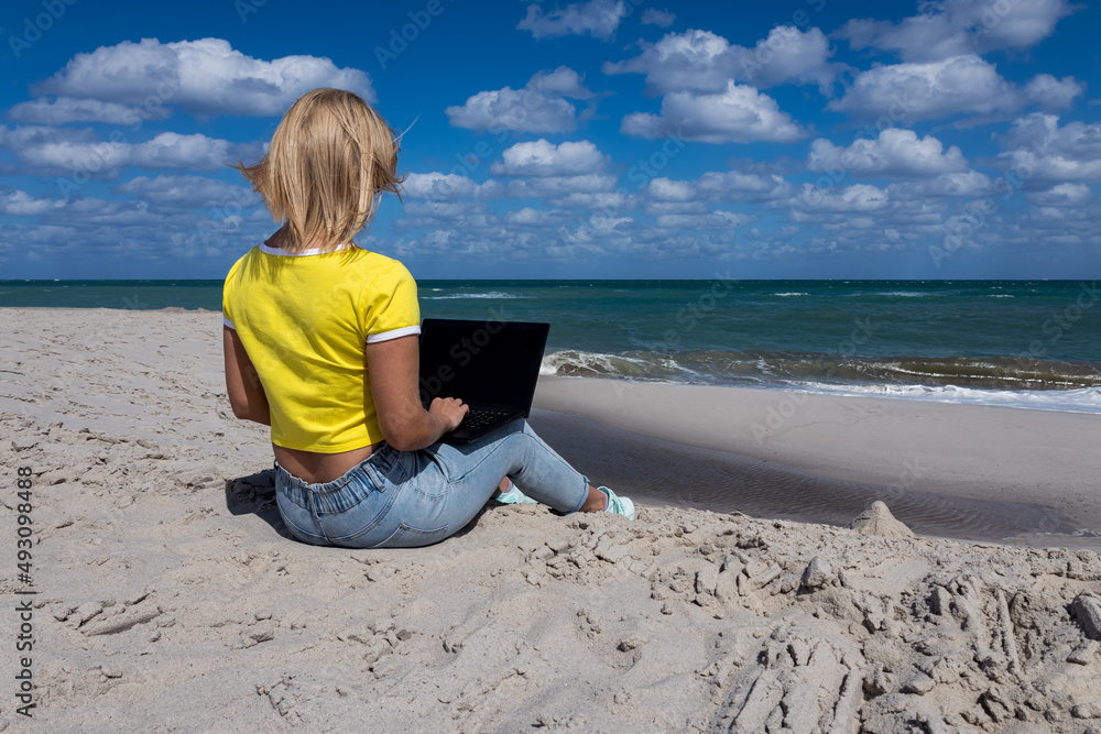 A blonde girl in a yellow T-shirt and jeans sits with a computer on the ocean. Vertical photo. Soft focus.