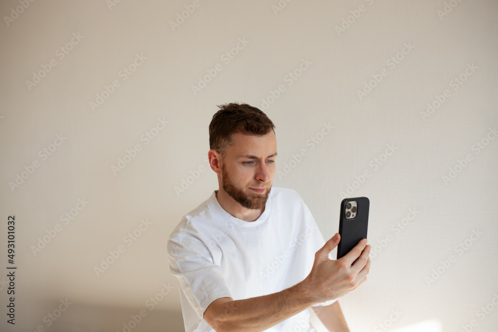 a young man with a beard 25-35 in a white t-shirt uses a smartphone a young man with a beard 25-35 in a white t-shirt uses a smartphone