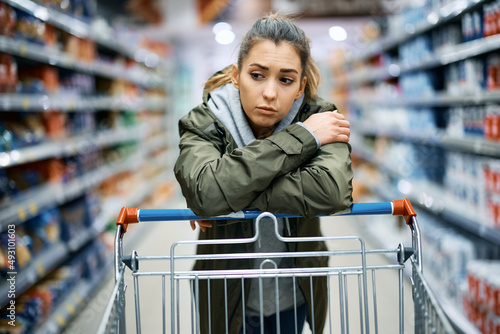 Young sad woman leans on shopping cart while standing among produce aisle at supermarket. photo