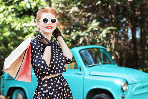 Beautiful stylish woman in vintage bright dotted clothes and glasses is holding shopping bags on background of mint old car. Fashion pin-up girl with makeup and hairstyle outdoor. Retro style concept. © Monstar Studio