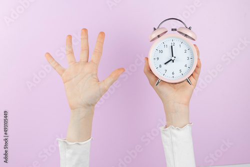Human arm is raised up holding pink alarm clock and showing open palm, five finger on pink background.