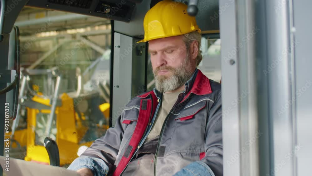Senior Caucasian technician in hardhat and workwear sitting in construction machine and using laptop while working at factory Senior Caucasian technician in hardhat and workwear sitting in construction machine and using laptop while working at factory