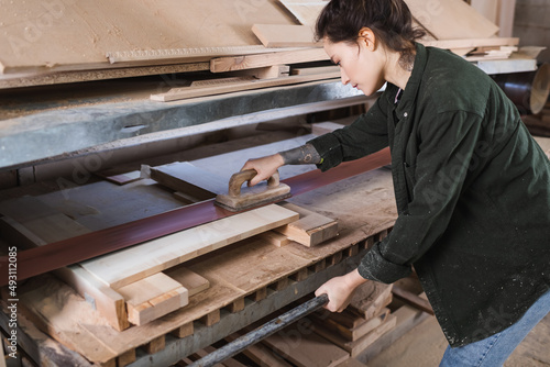 Side view of tattooed carpenter in sawdust polishing plank in workshop.