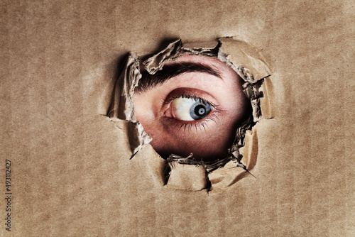 What is on the other side. Closeup portrait of an eye looking through a ripped hole in a piece of cardboard. photo