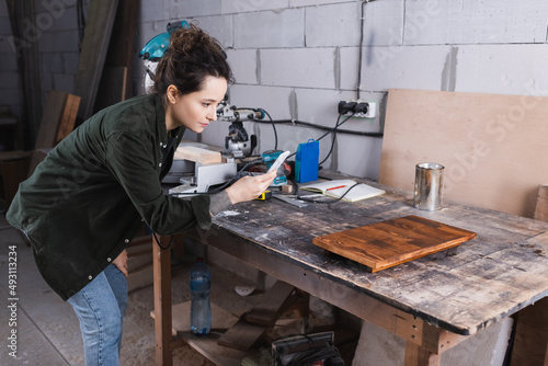Carpenter taking photo on smartphone near wooden board in workshop.