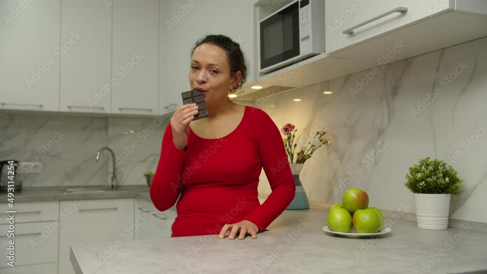 Beautiful mixed race woman enjoying taste of chocolate at home. Cheerful adult female showing satisfaction by eating sweets closing eyes in pleasure against background of domestic kitchen. Moving shot Beautiful mixed race woman enjoying taste of chocolate at home. Cheerful adult female showing satisfaction by eating sweets closing eyes in pleasure against background of domestic kitchen. Moving shot