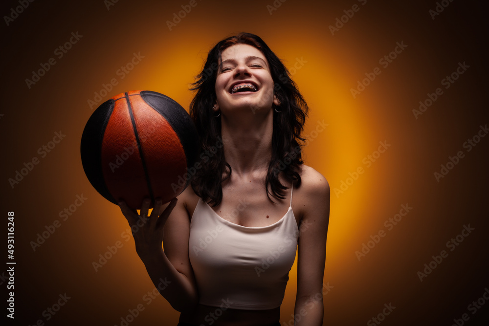 Teenage girl with dental braces holding basketball. Studio portrait on orange colored background..
