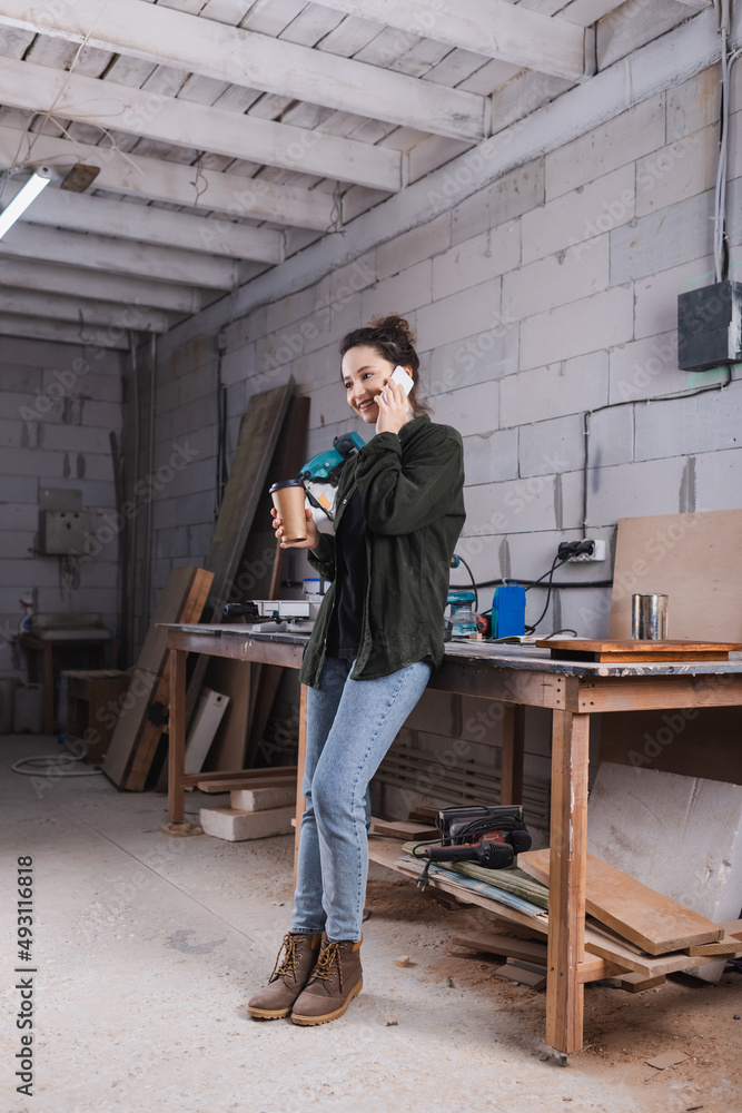 Smiling furniture designer holding coffee to go and talking on smartphone in workshop. Smiling furniture designer holding coffee to go and talking on smartphone in workshop.