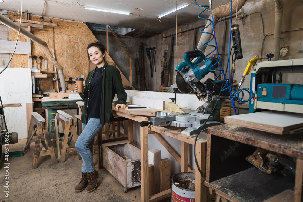 Smiling furniture designer holding coffee to go near gadgets and equipment in workshop. Smiling furniture designer holding coffee to go near gadgets and equipment in workshop.