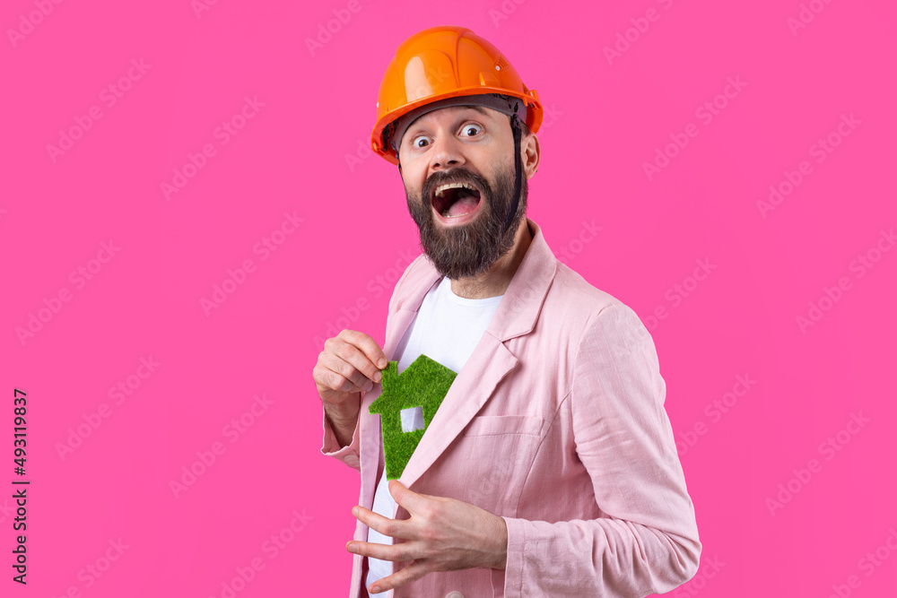 Portrait of young construction engineer wear orange hard hat, in a pink jacket standing on red studio background. A man holds a green eco house. Portrait of young construction engineer wear orange hard hat, in a pink jacket standing on red studio background. A man holds a green eco house.
