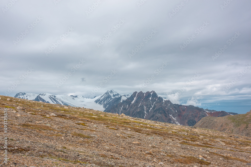 Dramatic landscape with sunlit diagonal stone mountainside with view to high snow mountains with sharp rocks and pointed peak under cloudy sky. Beautiful scenery with peaked top at changeable weather.