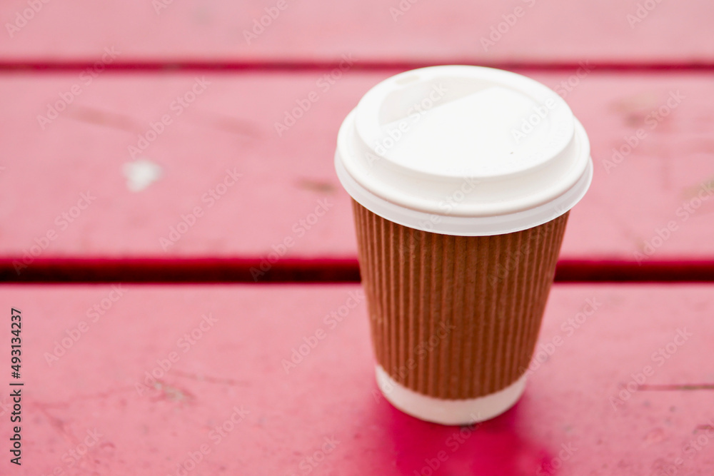 Cardboard cup with plastic lid for coffee or tee on a wooden red bench in a park. Outdoor refreshment concept. Selective focus. Cardboard cup with plastic lid for coffee or tee on a wooden red bench in a park. Outdoor refreshment concept. Selective focus.