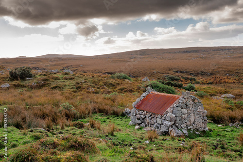 Small stone building with rusty red metal roof with beautiful nature scenery in the background. Connemara area, county Galway, Ireland. Storage hut in a field. Historical building. Cloudy sky