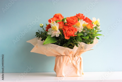 Fototapeta Naklejka Na Ścianę i Meble -  Beautiful bridal bouquet of different flowers alstromeria, small orange roses in cream colored wrapping paper stands on table on blue background. A gift for a woman, girl, mother, sister for March 8.