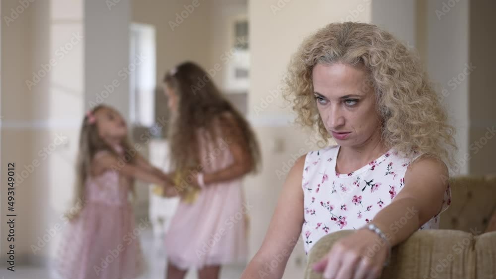 Overburdened Caucasian mother sighing rolling eyes in slow motion as daughters fighting for toy at background. Portrait of overwhelmed fatigue woman sitting at home with girls misbehave Overburdened Caucasian mother sighing rolling eyes in slow motion as daughters fighting for toy at background. Portrait of overwhelmed fatigue woman sitting at home with girls misbehave