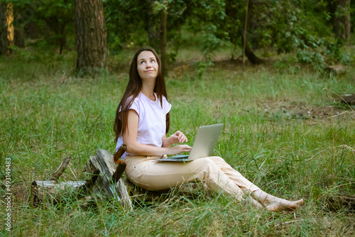 Dreamy beautiful young woman, girl with long brown hair sitting in a clearing in a woods, green forest, working at laptop. Remote work, study. Student with a computer in nature. Internet. Stay online.