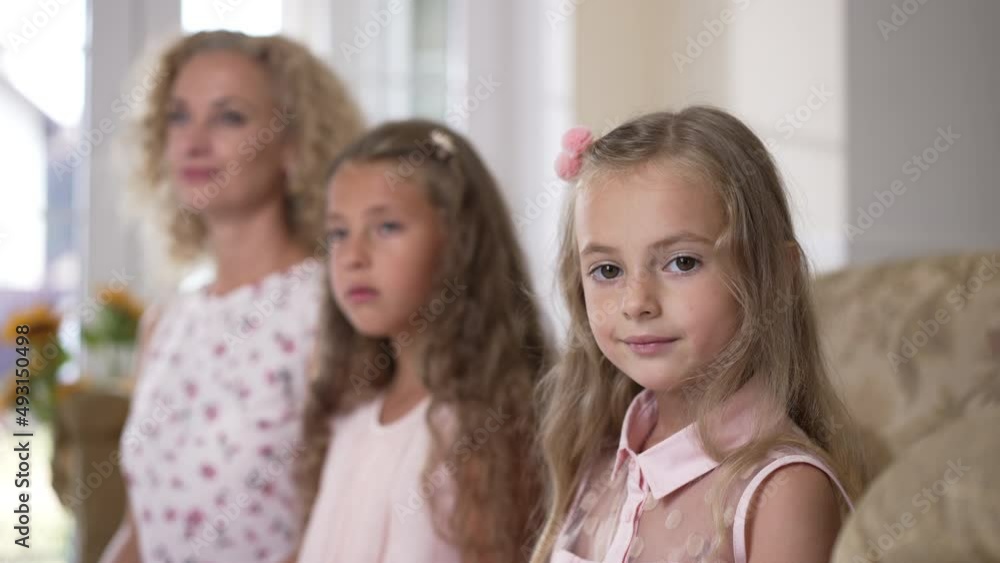 Cute girl looking at camera posing indoors with blurred sister and mother. Portrait of pretty Caucasian kid at home with family. Slow motion Cute girl looking at camera posing indoors with blurred sister and mother. Portrait of pretty Caucasian kid at home with family. Slow motion