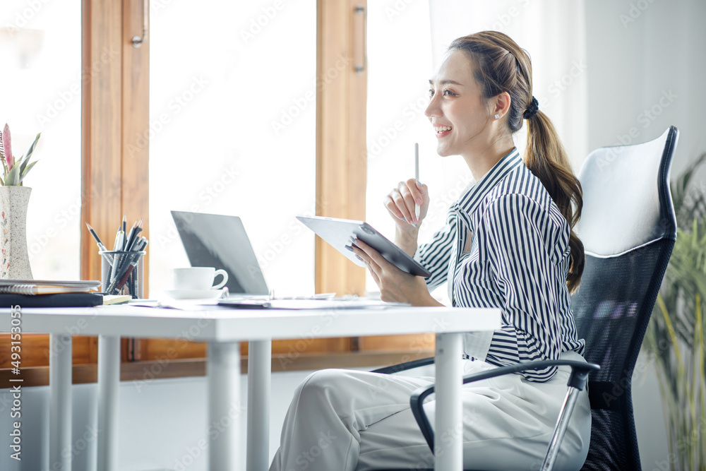 Smiling young Asian businesswoman working on a laptop computer at her desk in a bright modern home office, doing calculating expense financial report finance making notes on paper graph data document.