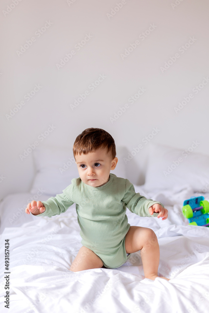 baby in a green cotton bodysuit is sitting on a white bed with a colored toy car. child is playing baby in a green cotton bodysuit is sitting on a white bed with a colored toy car. child is playing