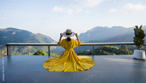 Asian female tourist in a beautiful yellow dress, holding a hat and sitting on the floor. With the beautiful sky and mountains of nature, Chiang Rai, Thailand.