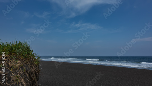 Turquoise waves of the Pacific Ocean roll onto the shore and foam. Tiny silhouettes of people on the black volcanic sand of the Khalaktyrsky beach. Blue sky. Green grass in the foreground. Kamchatka. photo