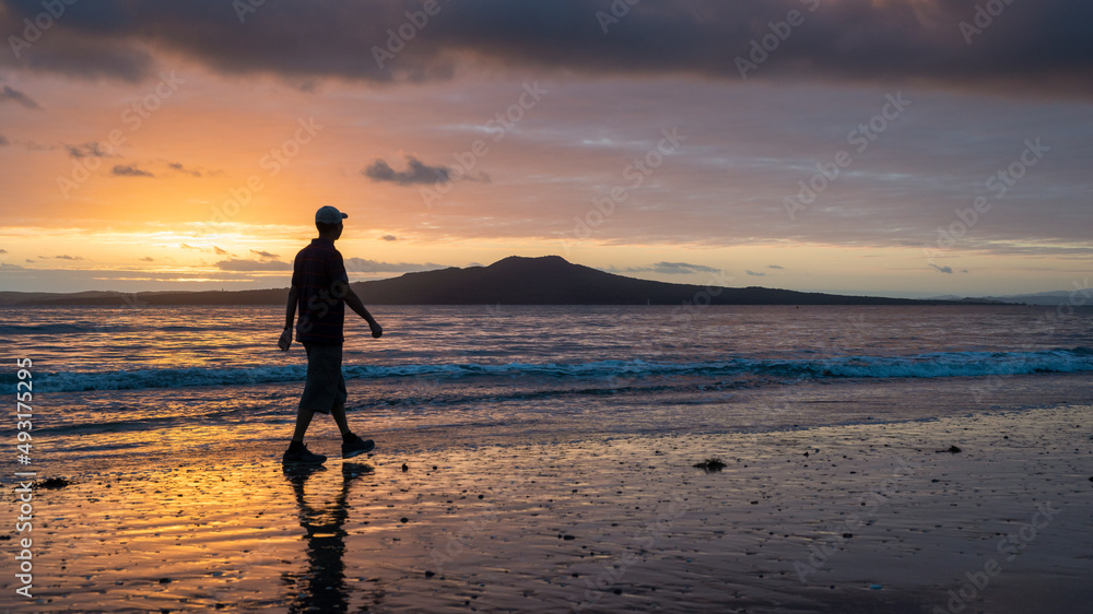 Silhouette man walking on Milford beach at sunrise, Rangitoto Island in the background, Auckland.
