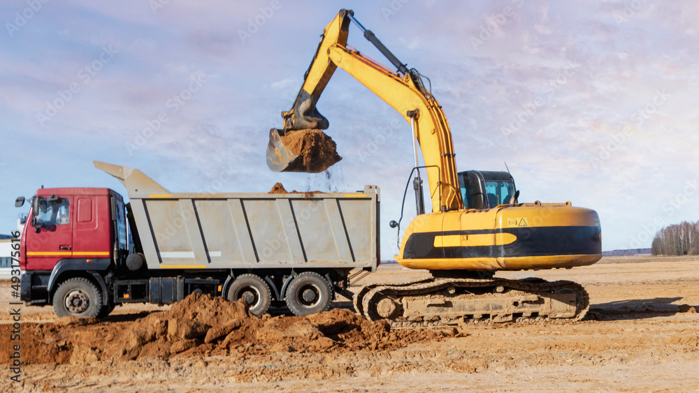 A powerful crawler excavator loads the earth into a dump truck against the blue sky. Development and removal of soil from the construction site. A powerful crawler excavator loads the earth into a dump truck against the blue sky. Development and removal of soil from the construction site.