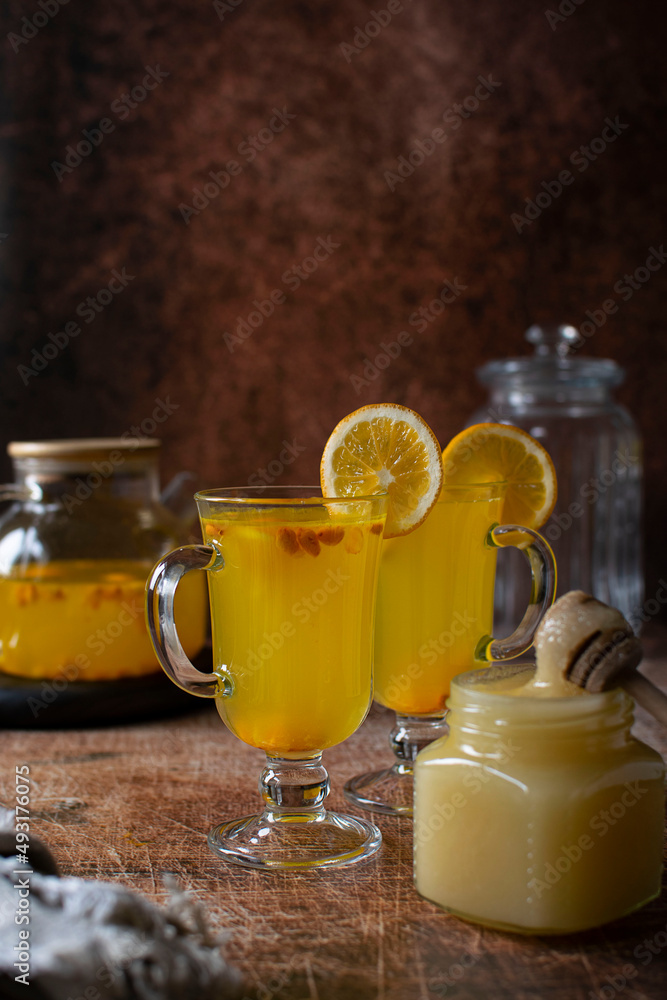 Fototapeta premium Two mugs with sea buckthorn drink, a teapot and honey on a wooden table. Image with copy space, vertical orientation, side view.