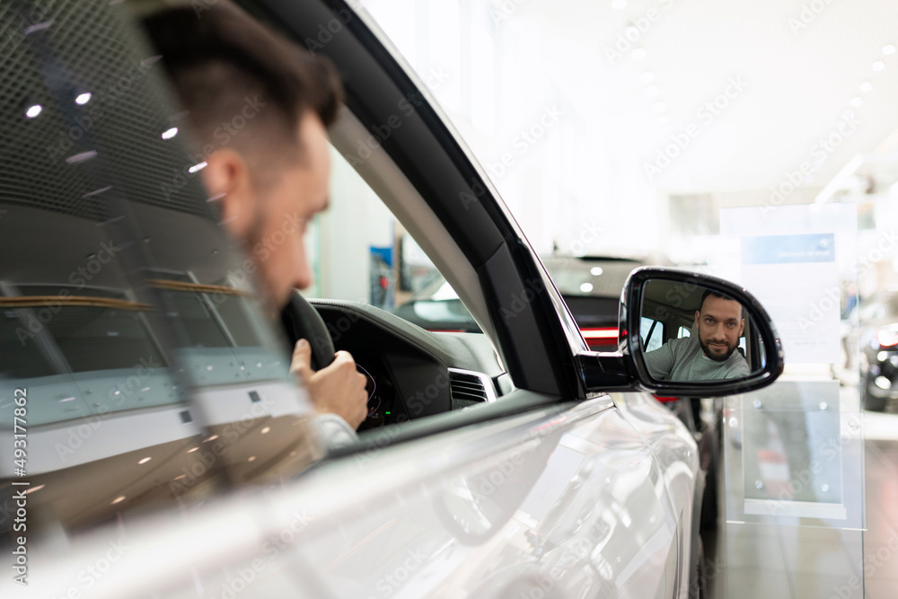 a man at a car dealership driving a car looks in the rearview mirror