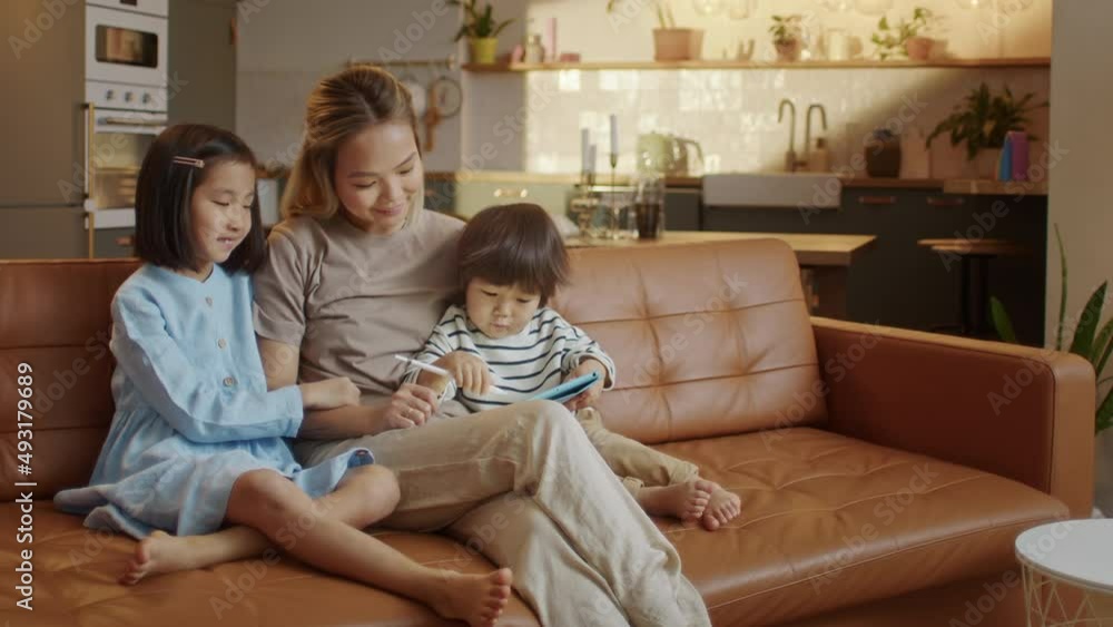 Vidéo Stock Loving Asian Mom and Little Daughter and Son Sit Together on Sofa at Home With Tablet Device Draw With Pencel, Using Education Application for Easy and Funny Learning, Modern Tech and Generation | Adobe Stock->