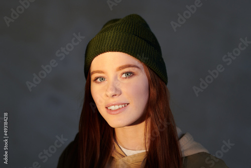 Horizonatal close-up shot of attractive young woman with darl red hair wearing cap smiling at camera, gray background photo