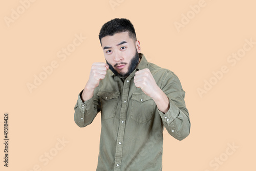 Boxing and self defense. Portrait of bearded asian man in casual clothes holding clenched fists, ready to defend himself in fight. isolated studio shot on beige background.