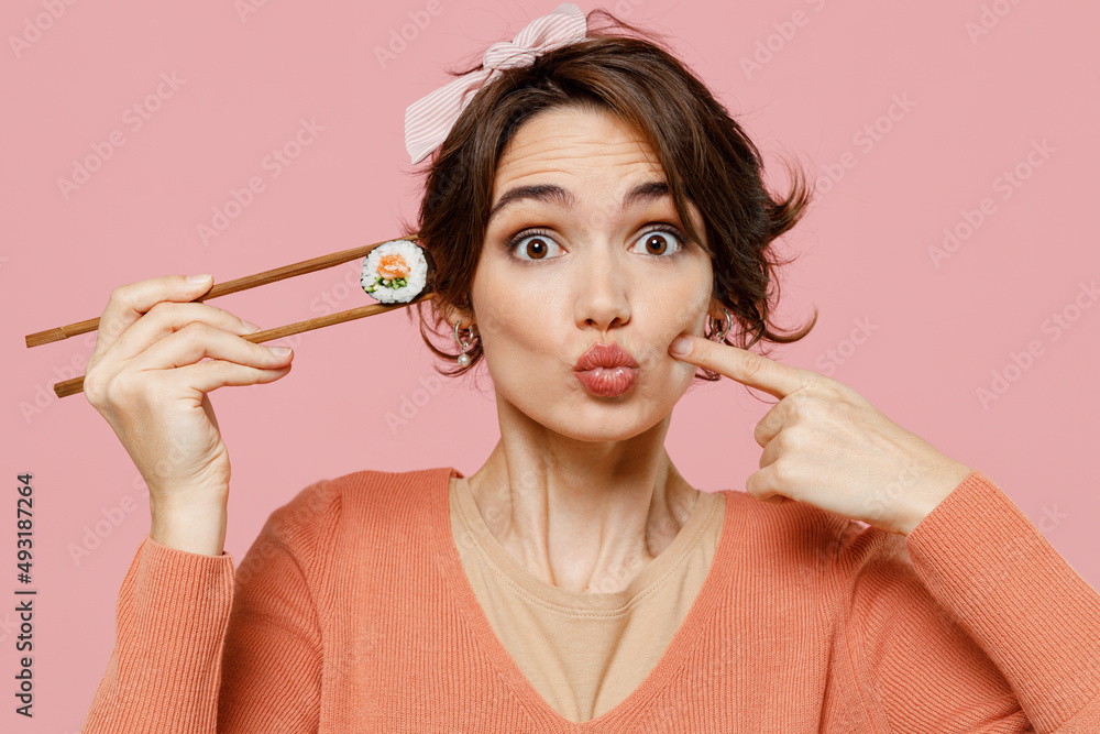 Young woman in sweater hold makizushi sushi roll served on black plate traditional japanese food point index fingers on blow puffing cheeks with funny face isolated on plain pastel pink background - obrazy, fototapety, plakaty 