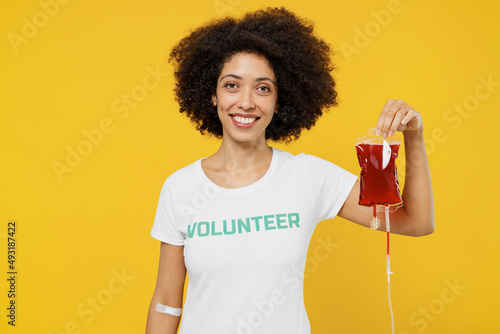 Фотография Young fun smiling happy donor woman of African American ethnicity wears white volunteer t-shirt hold bag with blood isolated on plain yellow background