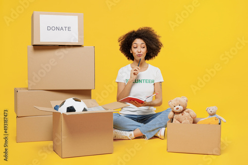 Full size minded young woman of African American ethnicity in white volunteer t-shirt sit near boxes with presents write down list isolated on plain yellow background Voluntary free work help concept photo