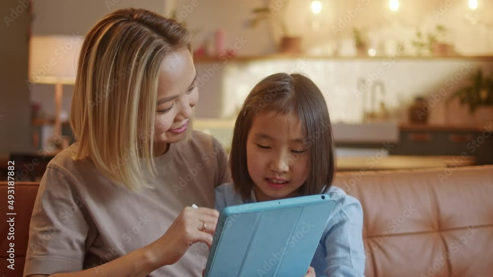 Asian Mom and Little Daughter Doing Homework by Using Tablet with Mother Help. Happy Smile Asia Kid While Sitting in the Living room at Night. Concept of Online Learning at Home. Asian Mom and Little Daughter Doing Homework by Using Tablet with Mother Help. Happy Smile Asia Kid While Sitting in the Living room at Night. Concept of Online Learning at Home.